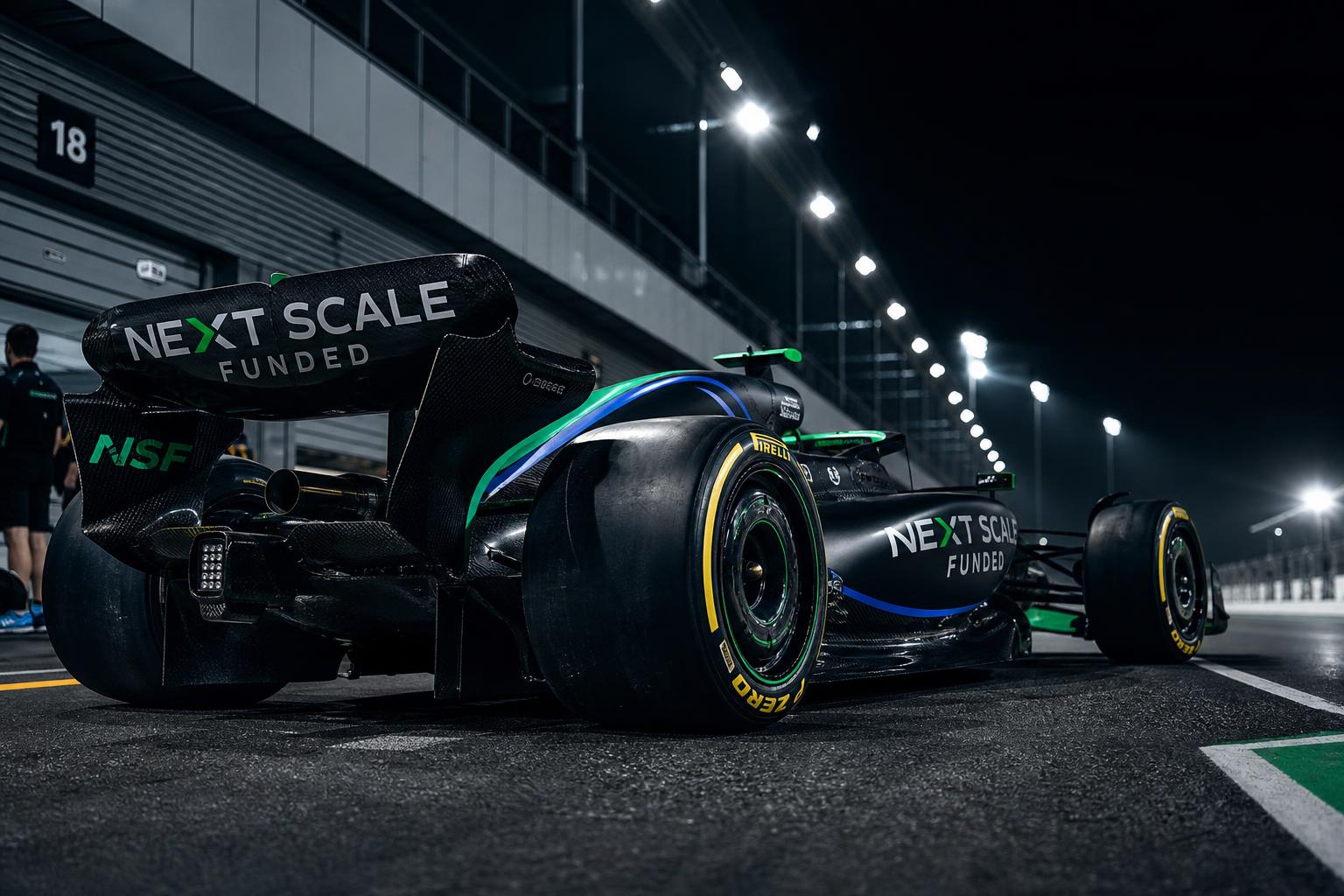 Formula 1 car in Next Scale Funded emerald and blue livery on a floodlit pit lane